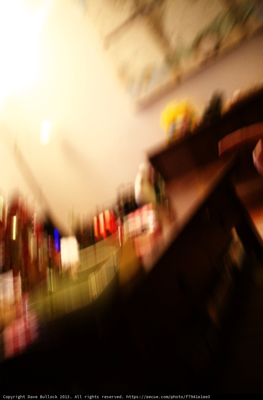 Blurry Finger on Plywood Surface Caption: A person holds a remote control against a background of plywood flooring, shelves, bottle, and furniture in an indoors location in Altadena, California. Tags include recreation, sport, and a pub environment.