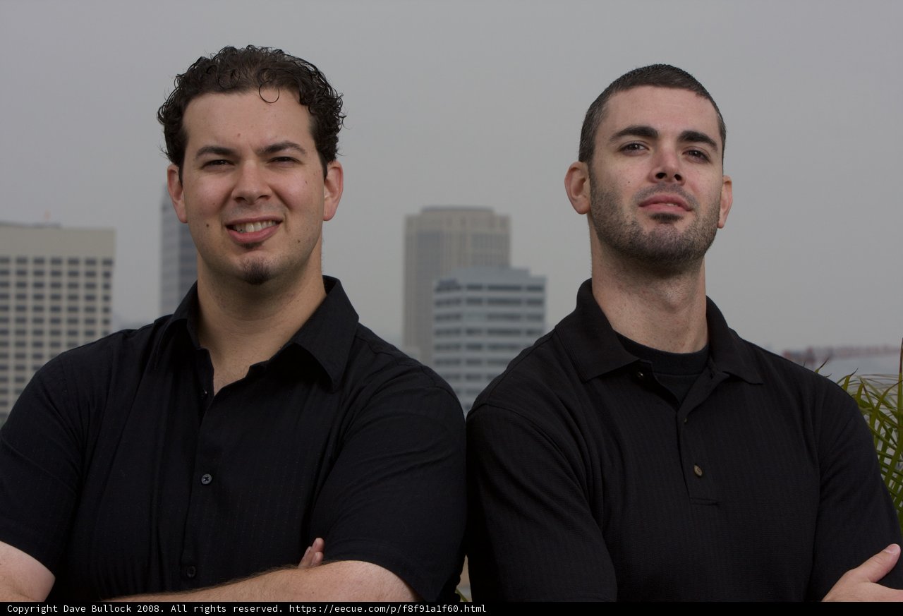Two Men with Black Shirts Pose in Front of Skyscraper