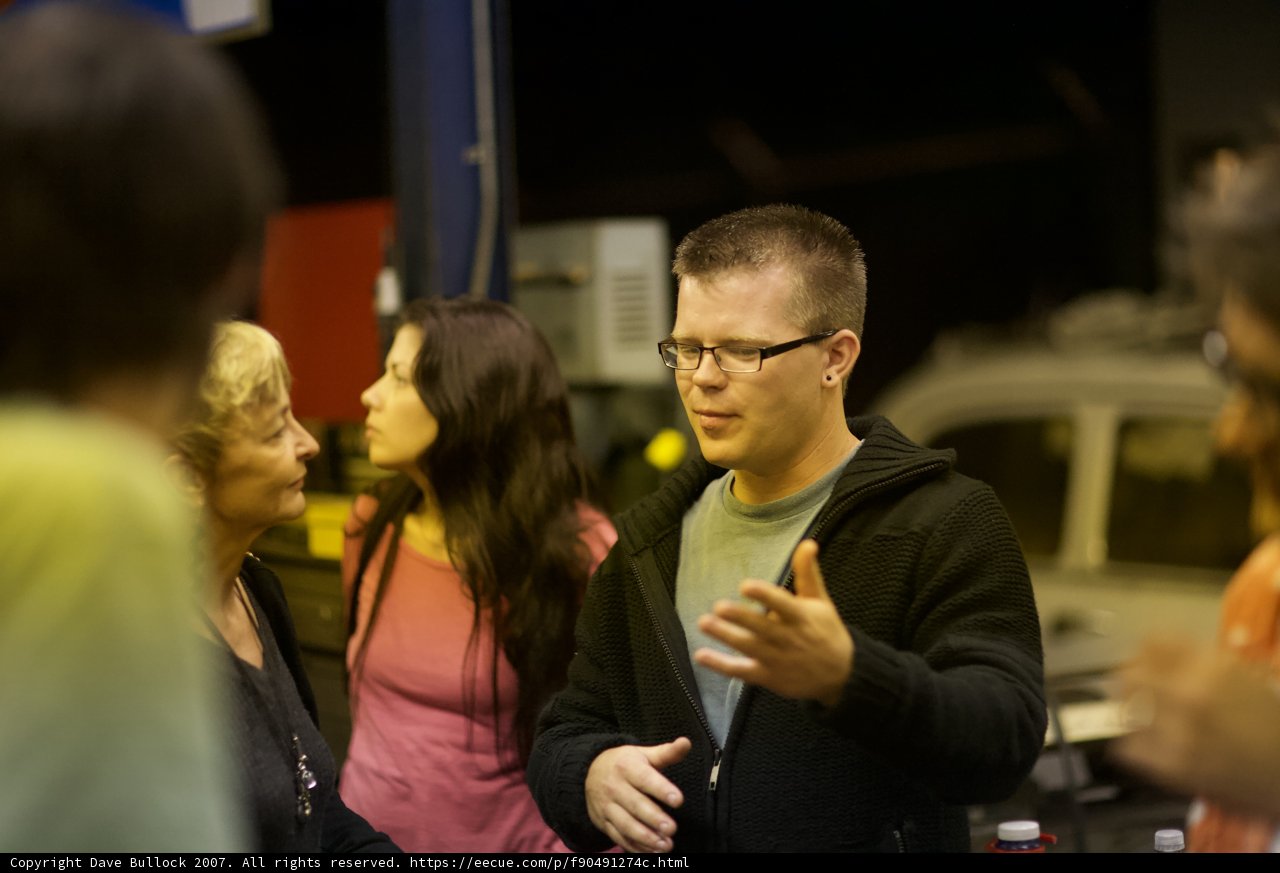 Garage Talk Caption: A man speaks to a group of five people inside a garage, adorned in eyeglasses and accessories, with fingers gesturing to emphasize his point. Two women and three adults listen attentively, all dressed in cozy knitwear and jewelry.