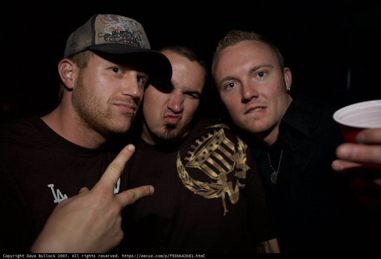 Three guys having a blast Caption: These three guys are having a great time at the party as they pose for a picture. Their hats and t-shirts display their love for baseball while they hold onto their cups and utensils.