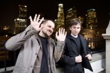 Two Men in Overcoats Pose in Front of Urban Metropolis