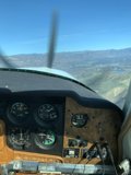 High in the Sky Caption: A close-up view of the cockpit dashboard and tachometer of a small airplane soaring over hills and land.