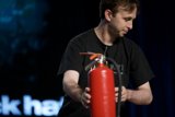 Ready to Fight Caption: A man stands with a fire extinguisher in hand, poised to battle any potential flames as he faces a screen full of people.
