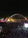 Nighttime Metropolis Crowd Caption: A large group of people gather in the heart of San Bernardino, surrounded by the bright lights of the city and the night sky, for a concert event.