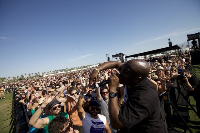 Shōgo Hamada Rocks Coachella Caption: Shōgo Hamada stands proud in front of a lively crowd at Coachella 2010. His all-black clothing and sunglasses add an aura of coolness to the concert atmosphere. The blue sky and fence surrounding the land add to the outdoor recreation vibe.