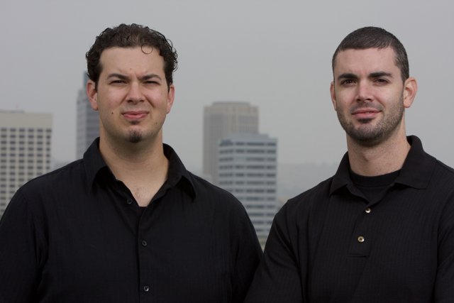 Two Men in Black Shirts Smile in Front of Skyscraper