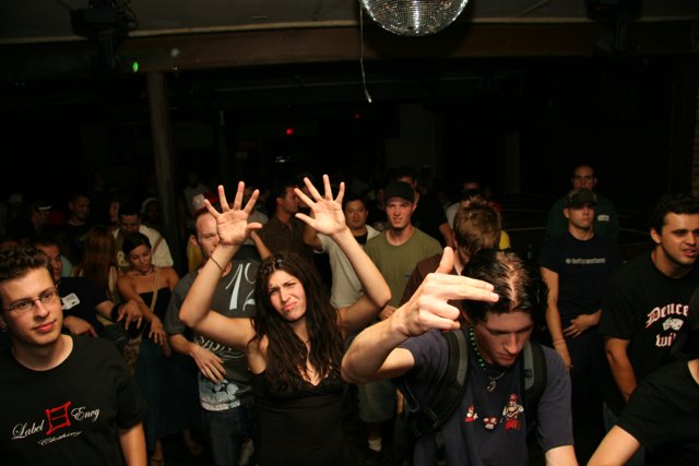 Black-Shirted Man in the Crowd Caption: This man stands out in the midst of a crowded nightclub, wearing a black shirt and accessorized with two necklaces and glasses.