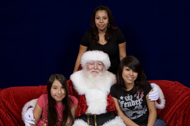 Santa Claus and the Merry Group Caption: Santa Claus poses with two girls and a woman at the 2008 APC Christmas party, all gathered comfortably on a couch.