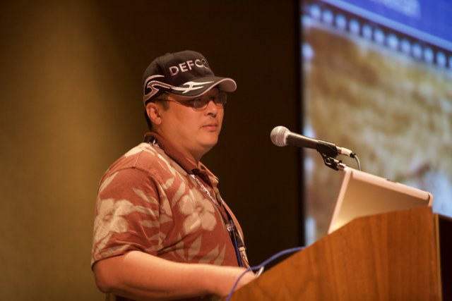 Yoshitomo Tani addresses the crowd Caption: Donning a baseball cap and sunglasses, Yoshitomo Tani delivers a speech at Defcon 17 while standing at the podium and speaking into a microphone.