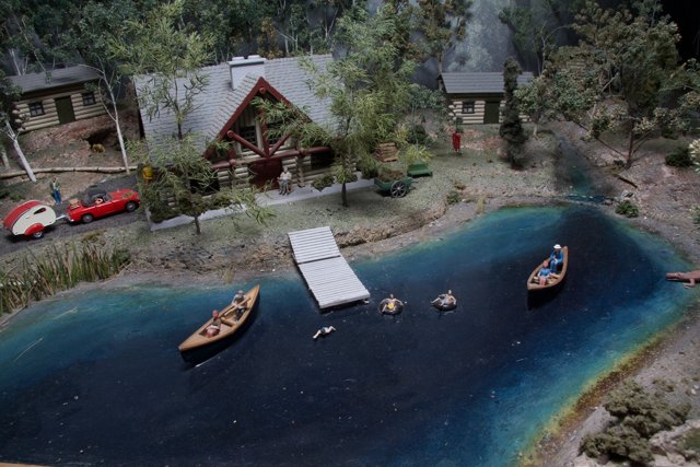 Lakeside House and Boats Caption: A miniature model of a cozy lakeside house with two boats docked in the water, surrounded by lush vegetation and bustling with activity with nine people enjoying various water sports and boat rides.