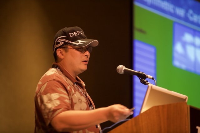Man addresses a crowded room wearing a hat and holding a microphone