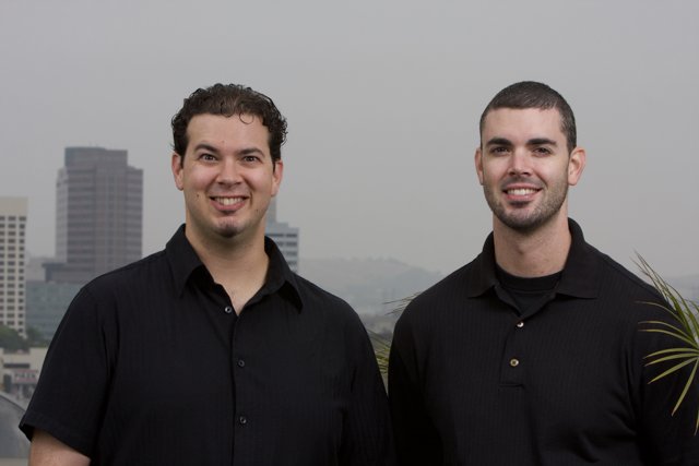 Two Smiling Men in Black Shirts Pose in Front of Cityscape