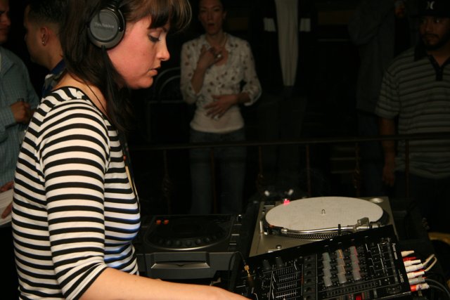 DJ Entertaining the Crowd Caption: A female deejay wearing headphones and standing behind a turntable entertains the crowd with her music at a club.