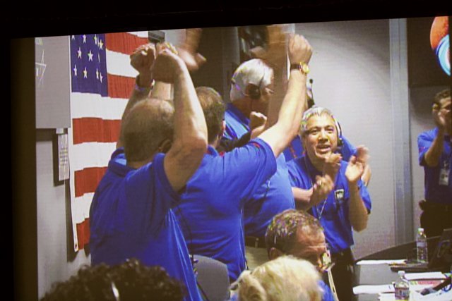 Celebrating Victory Caption: A group of 5 men wearing blue shirts cheer excitedly in front of a television displaying a football game at a bar, as one man holds up a flag in celebration.