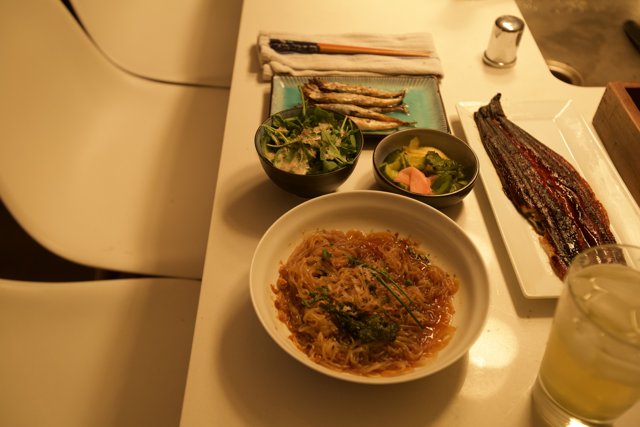 Delicious Noodle Dish on Wooden Table Caption: A savory bowl of vermicelli noodles and vegetables with a glass of water placed on a wooden table for a satisfying lunch.