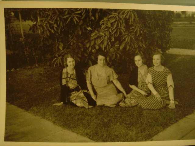 Garden Gathering Caption: Four women enjoy a peaceful afternoon sitting on the grass surrounded by lush foliage and beautiful plants.
