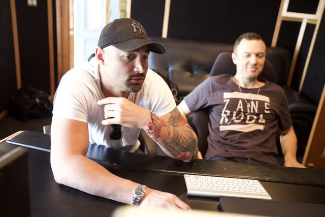 Recording Studio Session Caption: Chris Lake and a colleague sit at a desk in a recording studio in Altadena, California, surrounded by electronic equipment and music gear. Lake wears a baseball cap and a tattoo is visible on his arm, while his colleague works on a laptop.