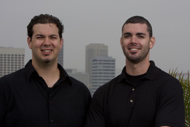 Two Smiling Men in Black Shirts Pose Next to Skyscraper