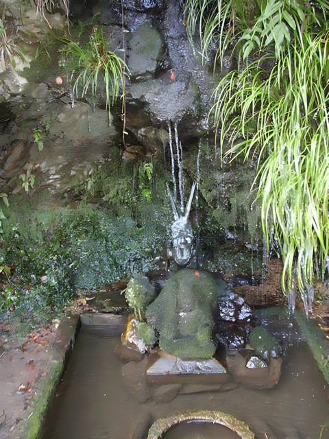 The Majestic Waterfall and Statue Oasis Caption: A serene natural haven showcasing the stunning beauty of flowing water and intricate statue in a lush green garden surrounded by moss and rocks.