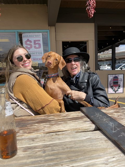 Two Women and a Dog Enjoying a Drink at a Wooden Table