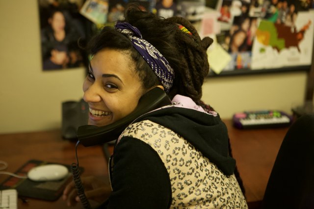 Dreadlocked Woman on the Phone Caption: Jodi G smiles while talking on the phone, surrounded by electronics and accessories at the APC office in 2008.