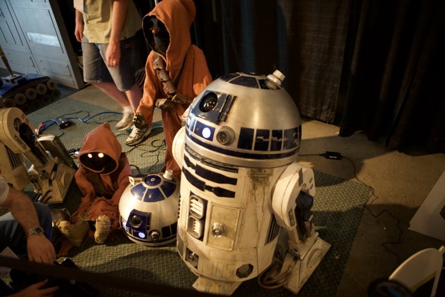 A Galactic Gathering Caption: Four people, including two babies, sit around a Star Wars R2D2 while wearing various clothing and headgear, including a helmet, and sporting two shoes and wristwatches.