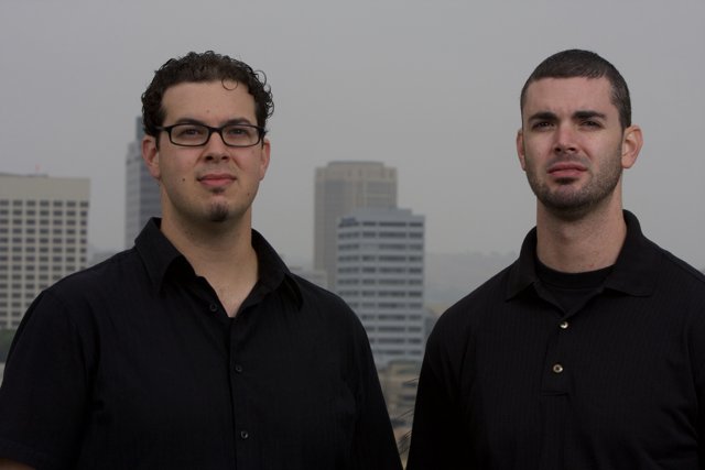 Two Smiling Men in Black Shirts Pose in Front of Skyscraper