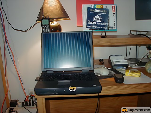 Working Hard Caption: A laptop computer on a desk covered in cords and surrounded by other electronics and furniture. A lamp shines down on the machine as it works diligently.