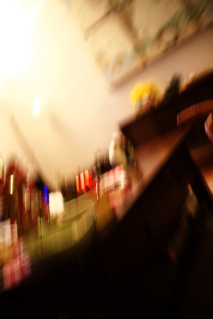Blurry Finger on Plywood Surface Caption: A person holds a remote control against a background of plywood flooring, shelves, bottle, and furniture in an indoors location in Altadena, California. Tags include recreation, sport, and a pub environment.