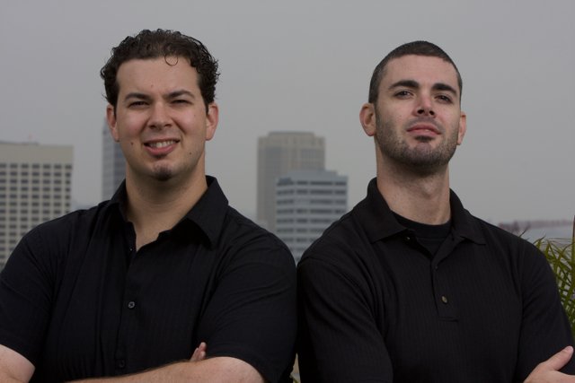 Two Men with Black Shirts Pose in Front of Skyscraper