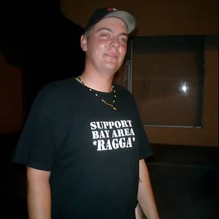Supporting Mike Taylor Caption: A portrait of a man wearing a black shirt and baseball hat with the words Support Mike Taylor while flaunting his accessories and a calm demeanor in nature.