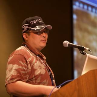Yoshitomo Tani addresses the crowd Caption: Donning a baseball cap and sunglasses, Yoshitomo Tani delivers a speech at Defcon 17 while standing at the podium and speaking into a microphone.