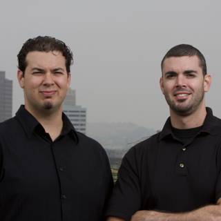 Two Smiling Men in Black Shirts with a Skyscraper in the Background
