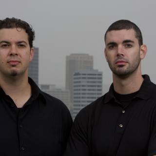 Two Men in Black Shirts Posing in Front of Skyscraper