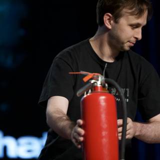 Ready to Fight Caption: A man stands with a fire extinguisher in hand, poised to battle any potential flames as he faces a screen full of people.
