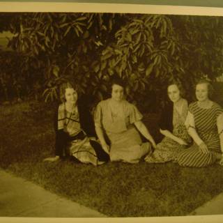 Garden Gathering Caption: Four women enjoy a peaceful afternoon sitting on the grass surrounded by lush foliage and beautiful plants.