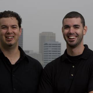 Smiling Duo in Black Shirts Pose Against a Skyscraper