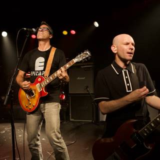 Two Guitarists Rocking the House Caption: Two talented men entertain a crowd with their guitars and microphones in a light-filled performance from the Bad Religion Glasshouse album in 2007.