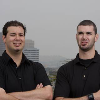 Two Smiling Men in Black Shirts Standing in a Cityscape