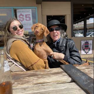 Two Women and a Dog Enjoying a Drink at a Wooden Table