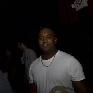 Commanding Attention Caption: A man stands confidently in front of a lively urban crowd at a night club wearing a T-Shirt and accessories, including a necklace and earbuds.