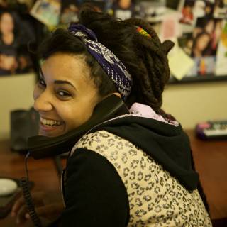 Dreadlocked Woman on the Phone Caption: Jodi G smiles while talking on the phone, surrounded by electronics and accessories at the APC office in 2008.