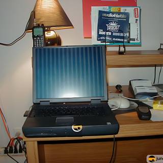 Working Hard Caption: A laptop computer on a desk covered in cords and surrounded by other electronics and furniture. A lamp shines down on the machine as it works diligently.