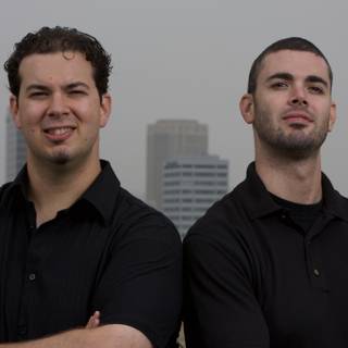 Two Men with Black Shirts Pose in Front of Skyscraper